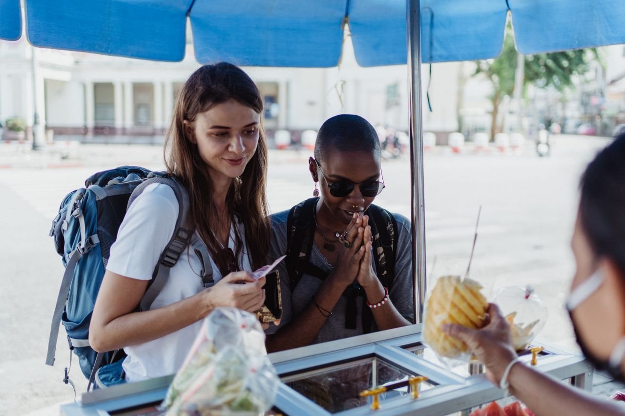 Two women travelers purchase street food from a vendor, enjoying a cultural experience outdoors.