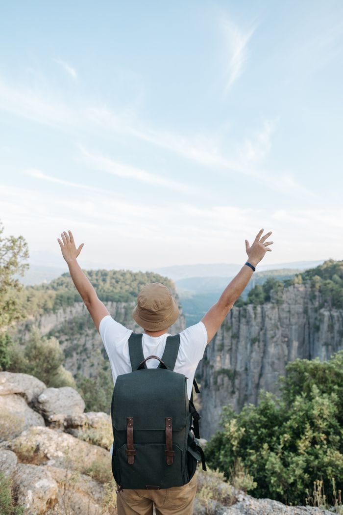 Man with arms raised celebrates nature on cliff edge overlooking a canyon.